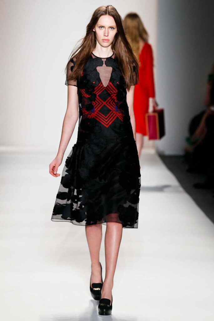 Model walks runway in a black and red dress with sheer details and black heels on a white catwalk.
