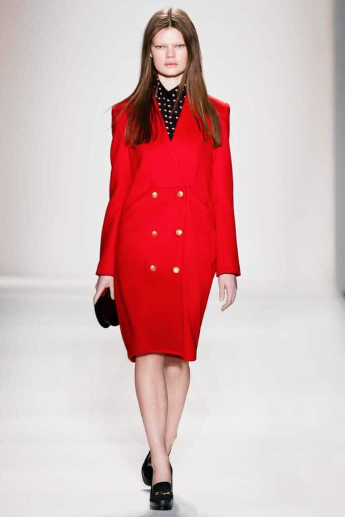Woman on runway wears a red double-breasted coat with gold buttons and a black polka dot blouse.