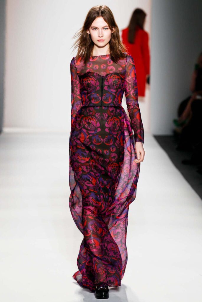 Model walks runway in a sheer, long-sleeved, purple and red patterned dress with flowing fabric.