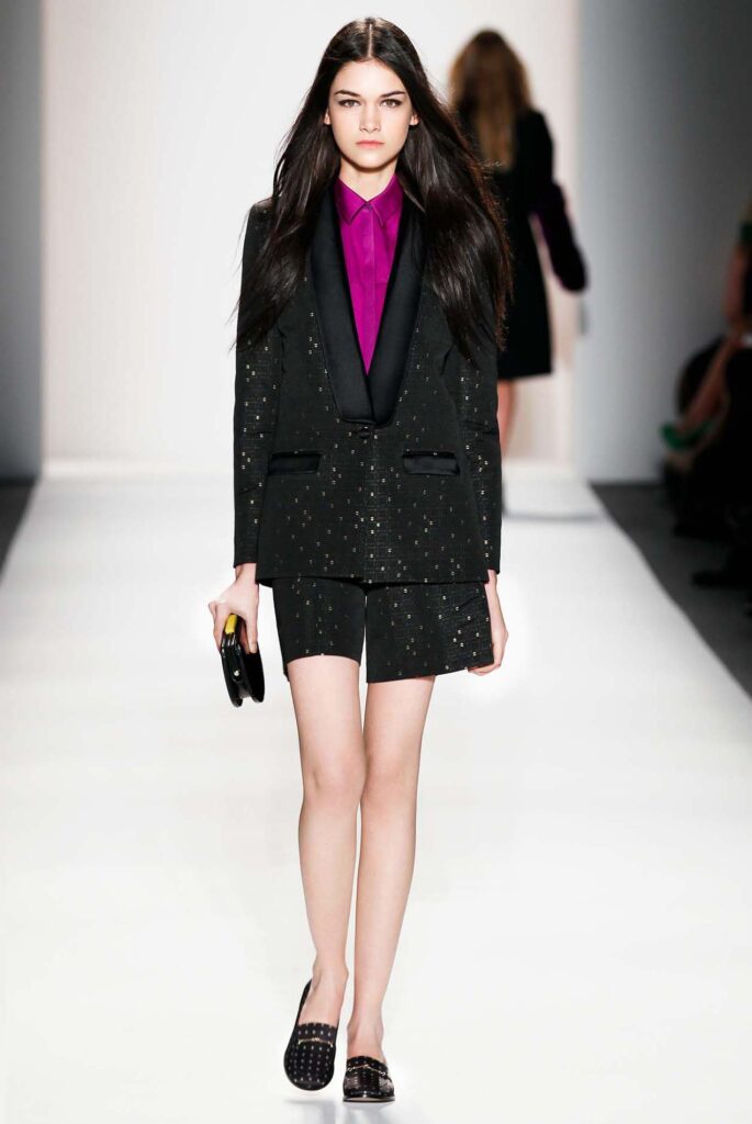 Model wearing a black tailored shorts suit with a bright magenta shirt walking on a fashion runway.