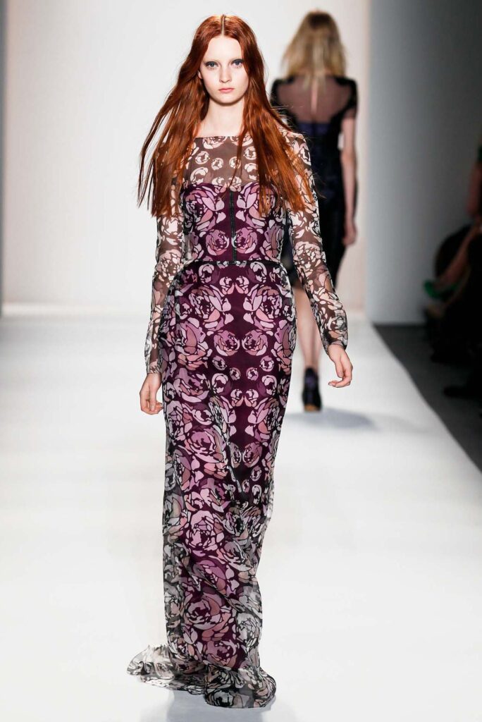 Model in a sheer, long-sleeved floral gown walks a white runway at a fashion show.