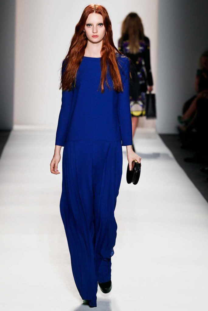 Model in a flowing royal blue outfit walks down a runway, carrying a small black clutch.