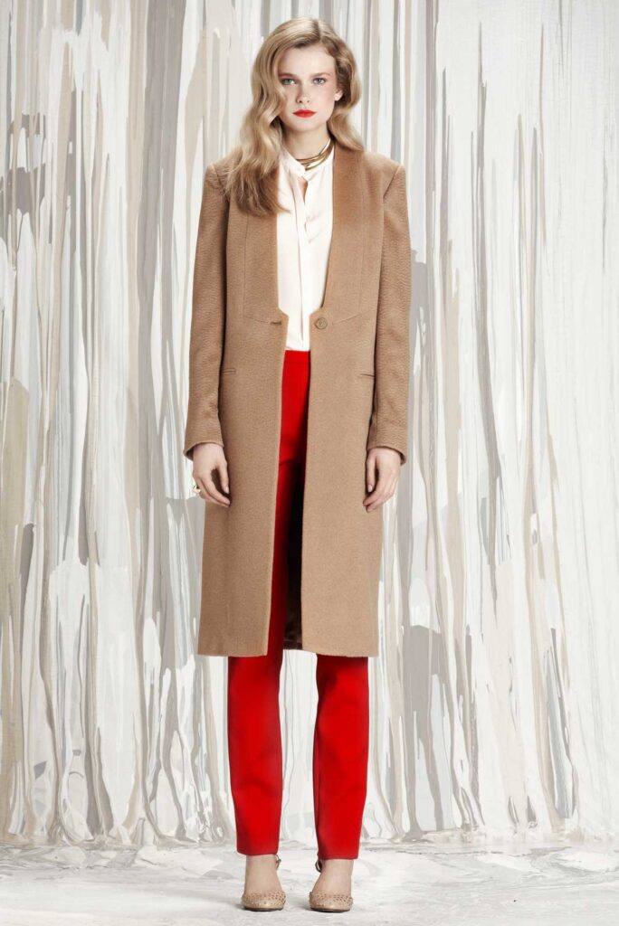 Woman in tan long coat, white blouse, red pants, and heels stands in front of abstract striped curtains.