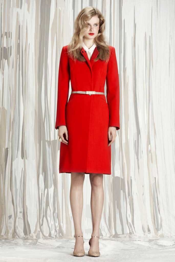 Woman in a red belted coat, beige heels, and red lipstick stands against a light, abstract background.