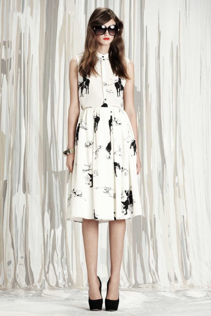 Woman in a sleeveless white dress with black animal print, wearing sunglasses and black heels, standing indoors.