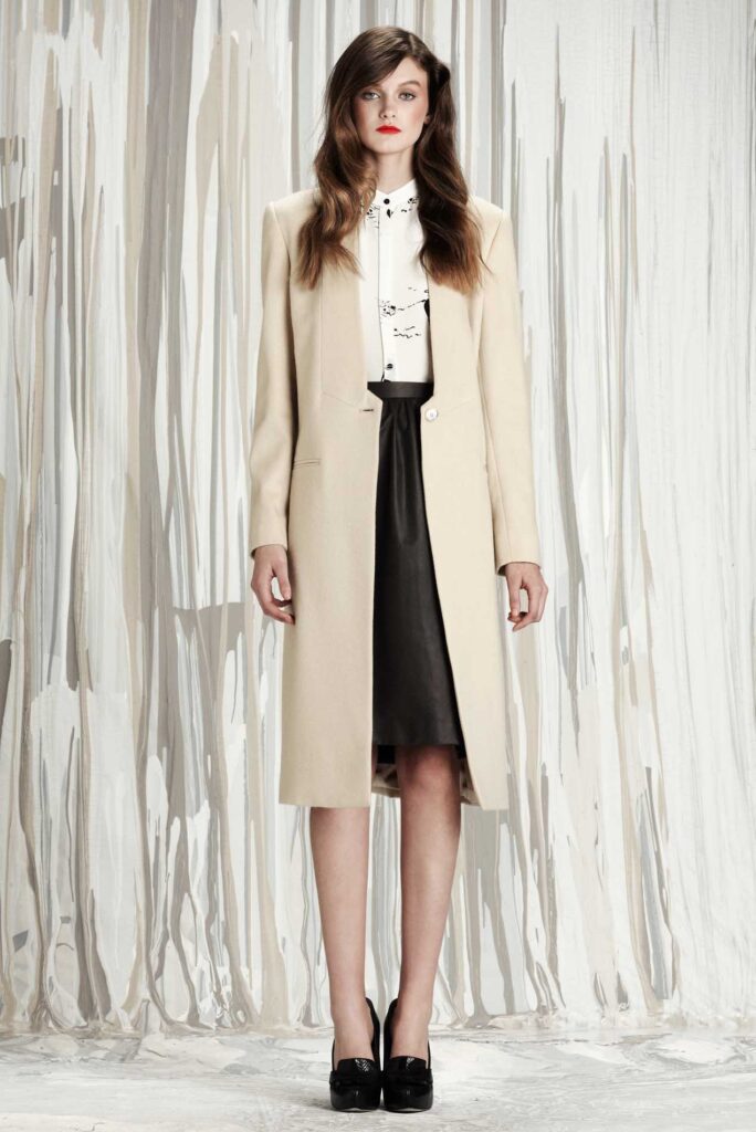 Woman in beige coat, white blouse, black skirt, and black heels standing in front of a gray curtain backdrop.