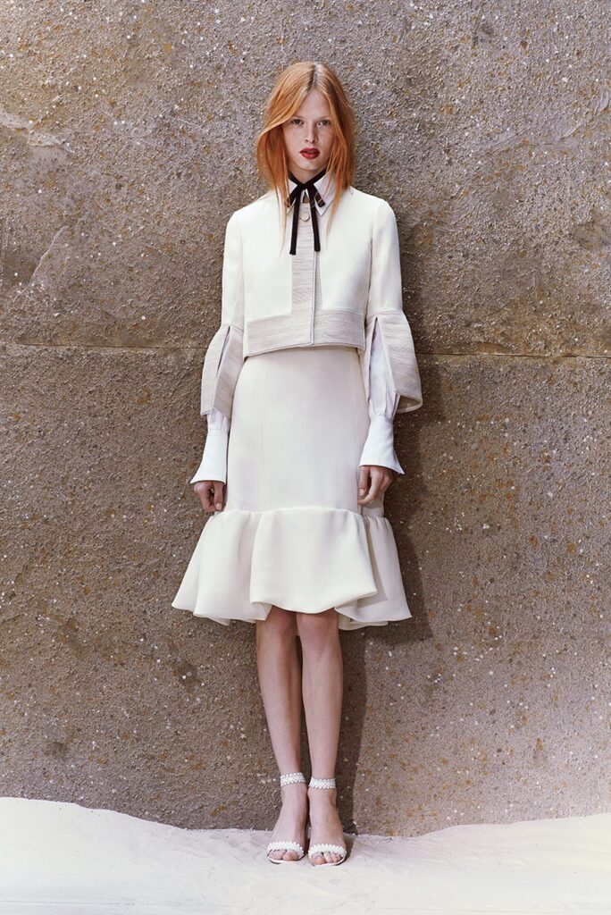 Woman in a white ruffled dress and jacket stands against a textured stone wall, looking at the camera.