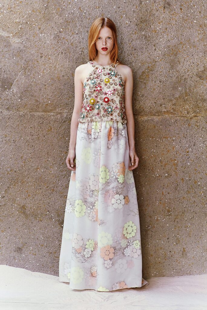 Woman in a sleeveless floral top and a long pastel flower-patterned skirt stands against a textured wall.