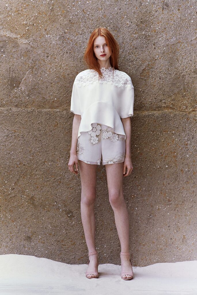 Woman with long red hair wearing a white top and shorts stands against a textured beige wall.
