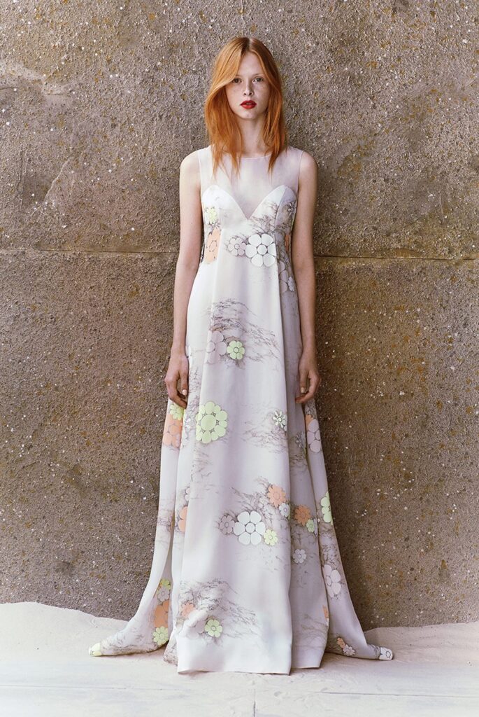 Woman in a long, sleeveless floral dress stands against a textured stone wall, looking directly ahead.