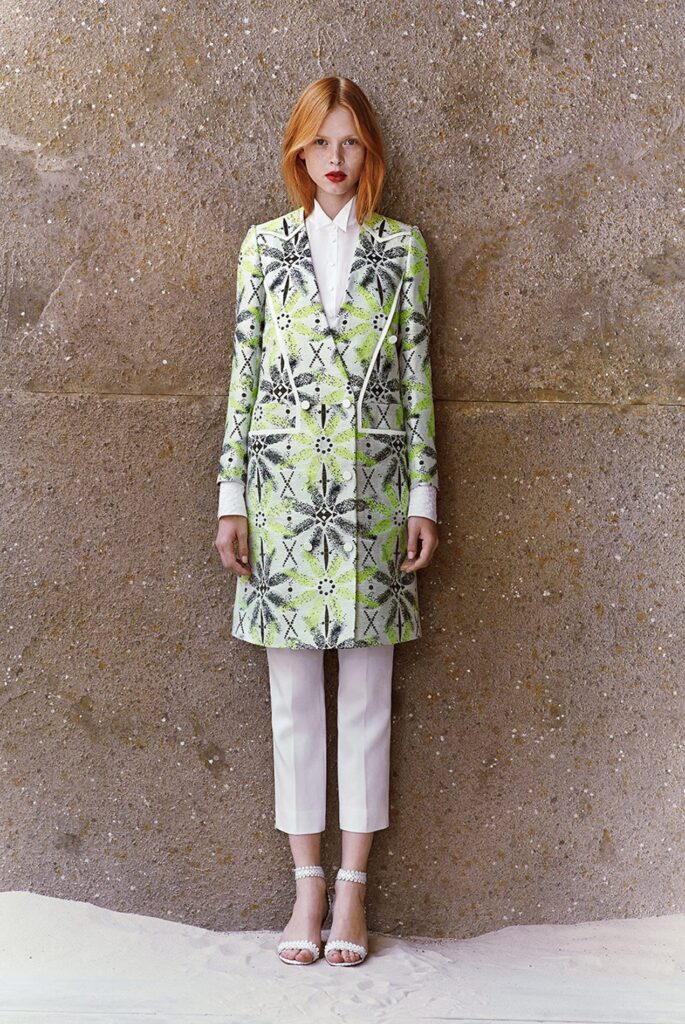 Woman in patterned green coat, white pants, and sandals stands against a textured, sandy wall.