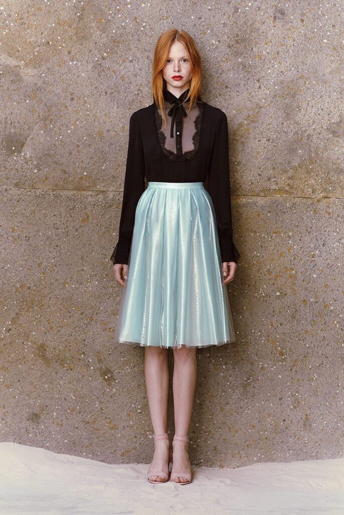 Woman in sheer black blouse with bow and shiny light blue skirt standing against a textured wall.
