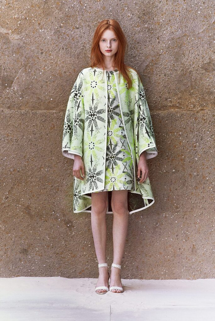 Woman with long red hair wears a green patterned dress and matching cape, standing against a stone wall.