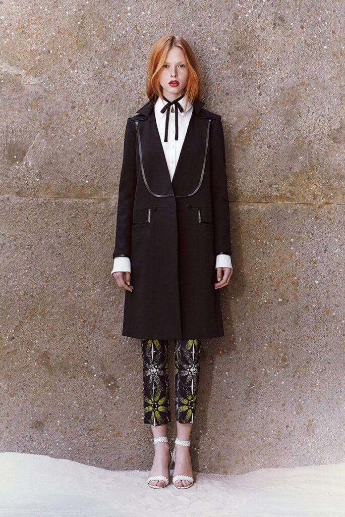 A woman in a long black coat, patterned pants, and heels stands against a textured wall.