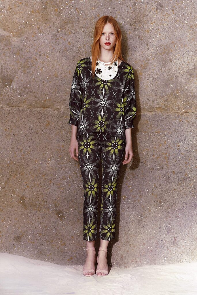 Woman in patterned black and yellow pants and top, standing against a textured wall, wearing pink heels.