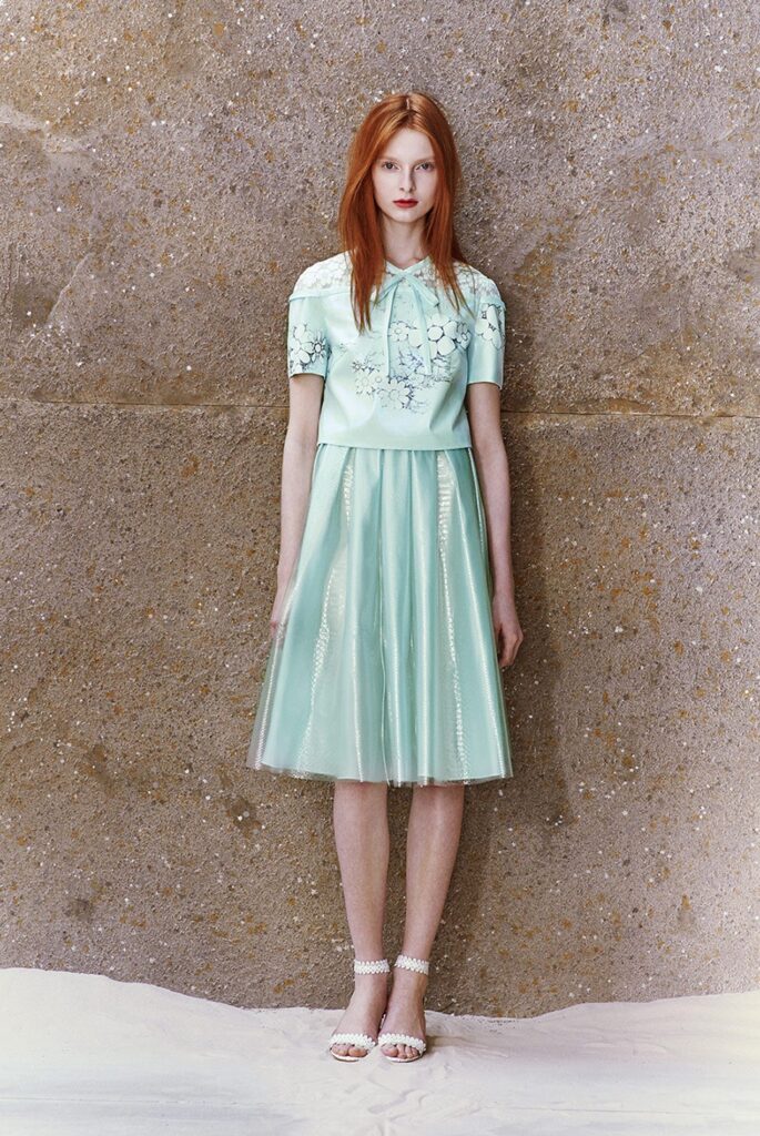 Woman with red hair in a mint green dress and white heels standing against a textured wall.