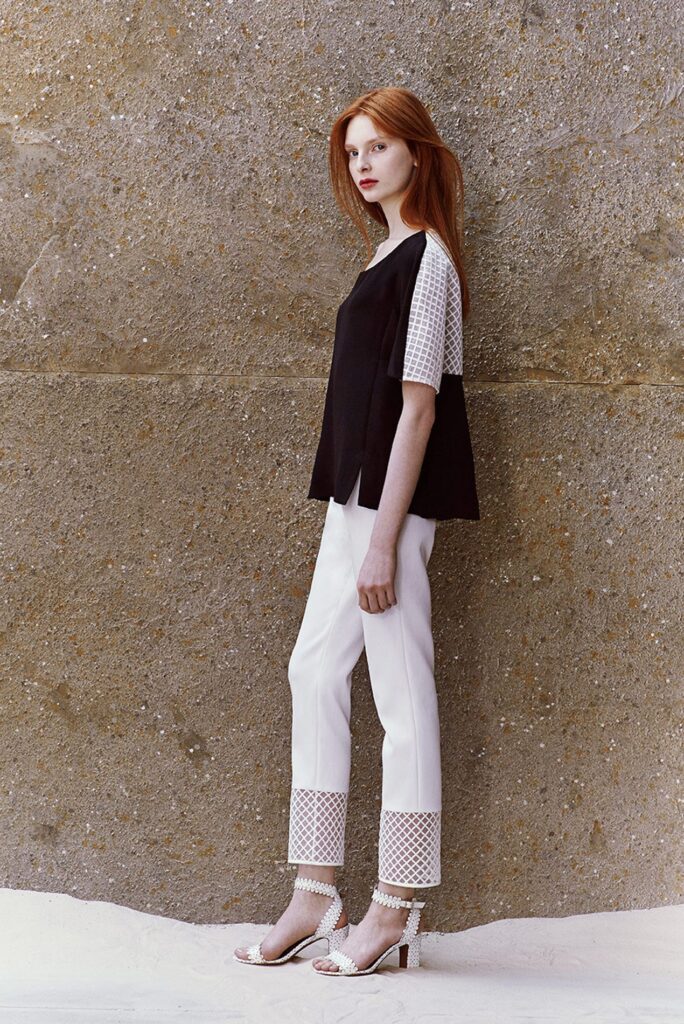 Woman in black top with white sleeves, white pants with mesh cuffs, and white heels stands by a stone wall.