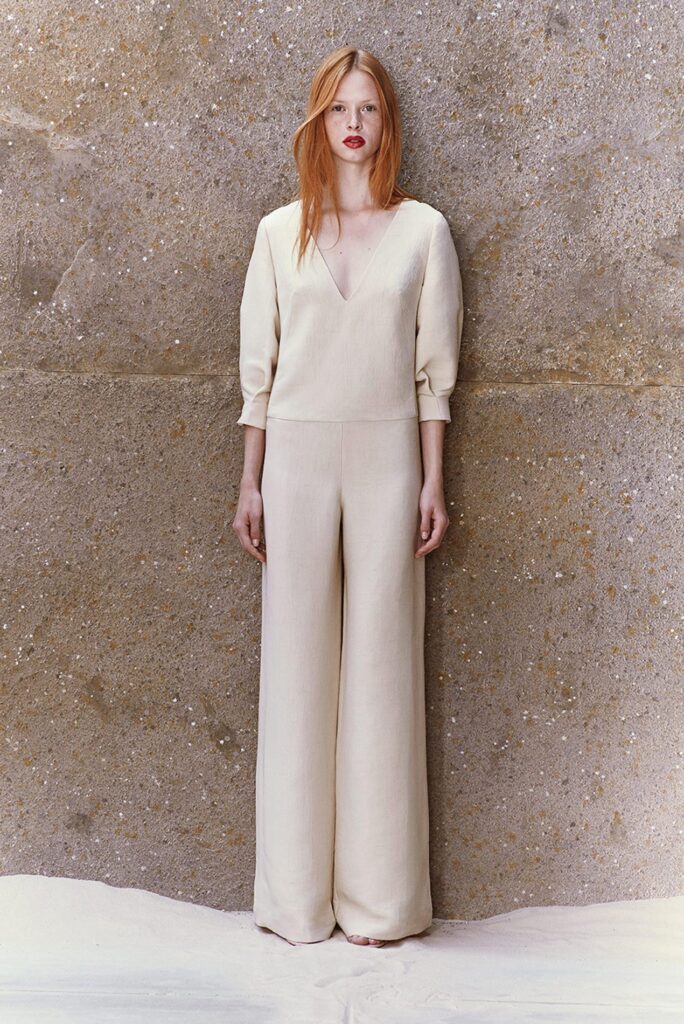 Woman with long red hair in a cream jumpsuit stands against a textured brown wall with sand at her feet.