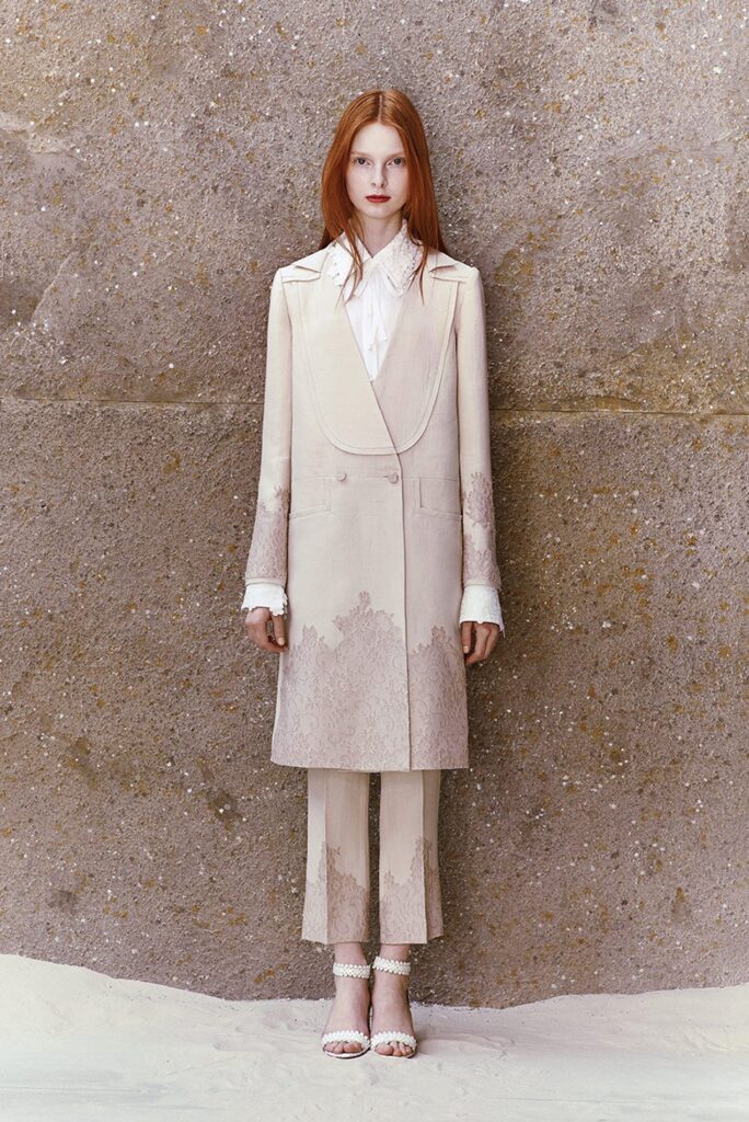 A woman in a cream-colored suit stands against a textured wall, looking straight at the camera.