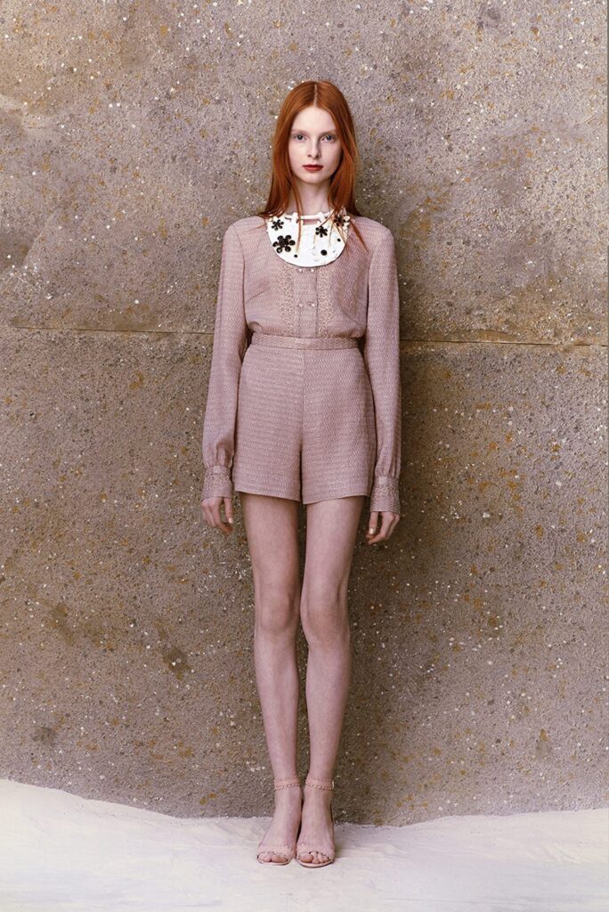 Woman with long red hair wears a beige outfit with shorts and a white, flowered collar, standing by a textured wall.