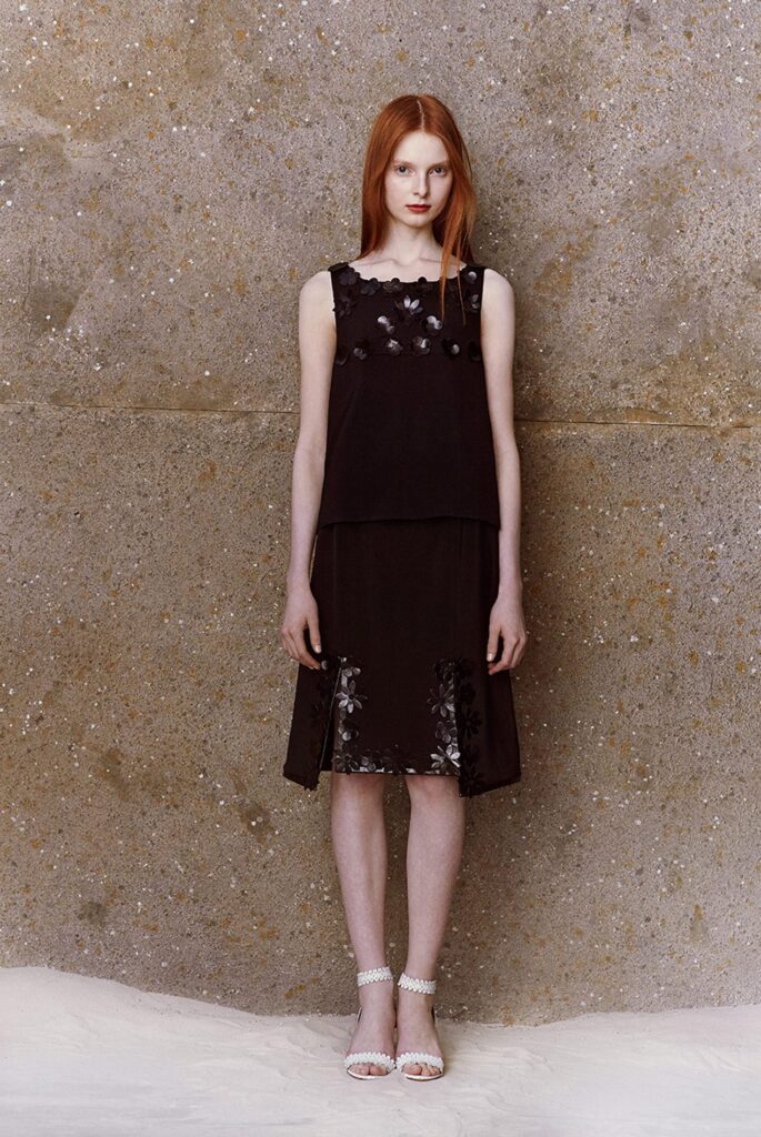 Woman with long red hair in a sleeveless black dress, standing against a textured, stone-like wall.
