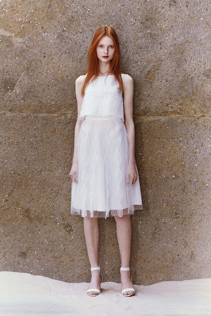 Woman with long red hair wears a sleeveless, knee-length white lace dress and white heels, standing by a wall.