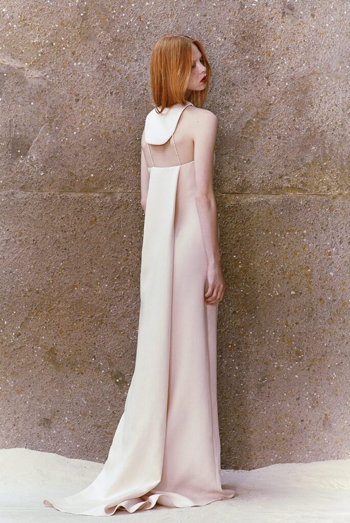 Woman with long red hair in a pale pink backless gown stands on sand against a textured stone wall.