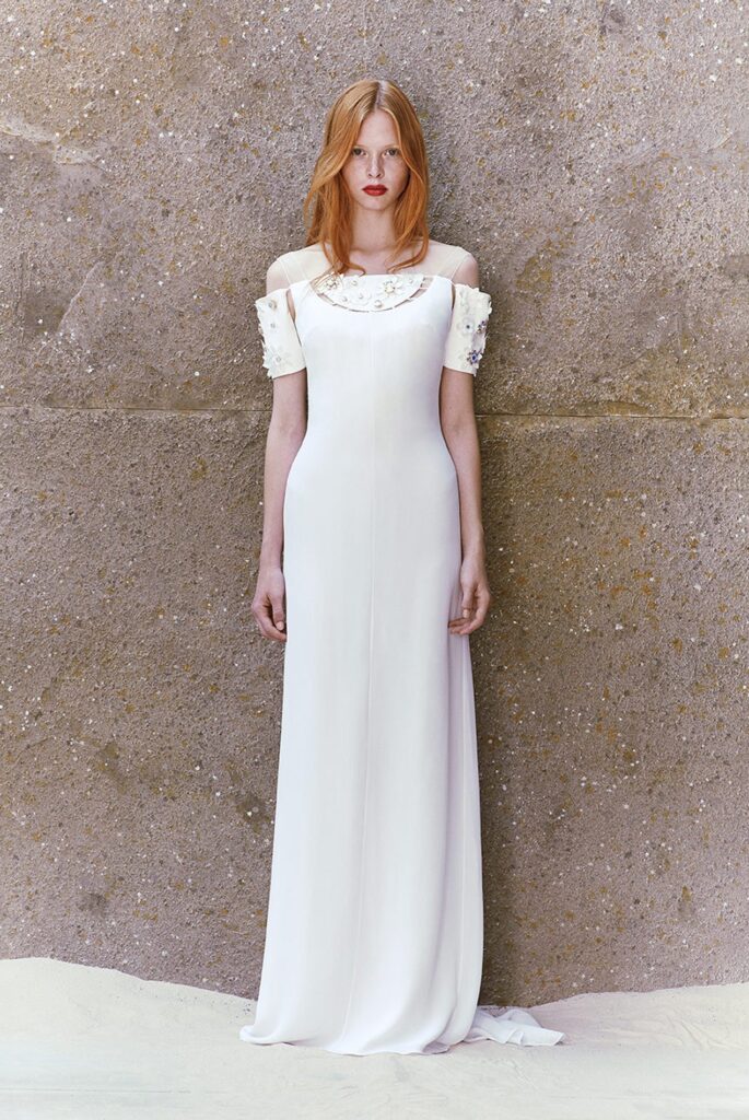 Woman in a white off-shoulder gown with embellishments, standing against a textured stone wall.
