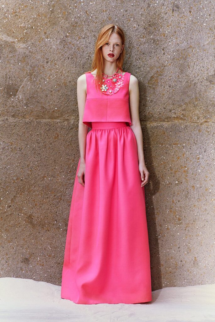 Woman in a sleeveless bright pink gown with floral embellishments, standing against a textured wall.
