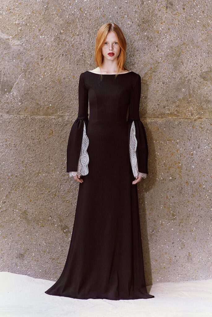 Woman in a long black dress with flared sleeves and lace details, standing against a textured stone wall.