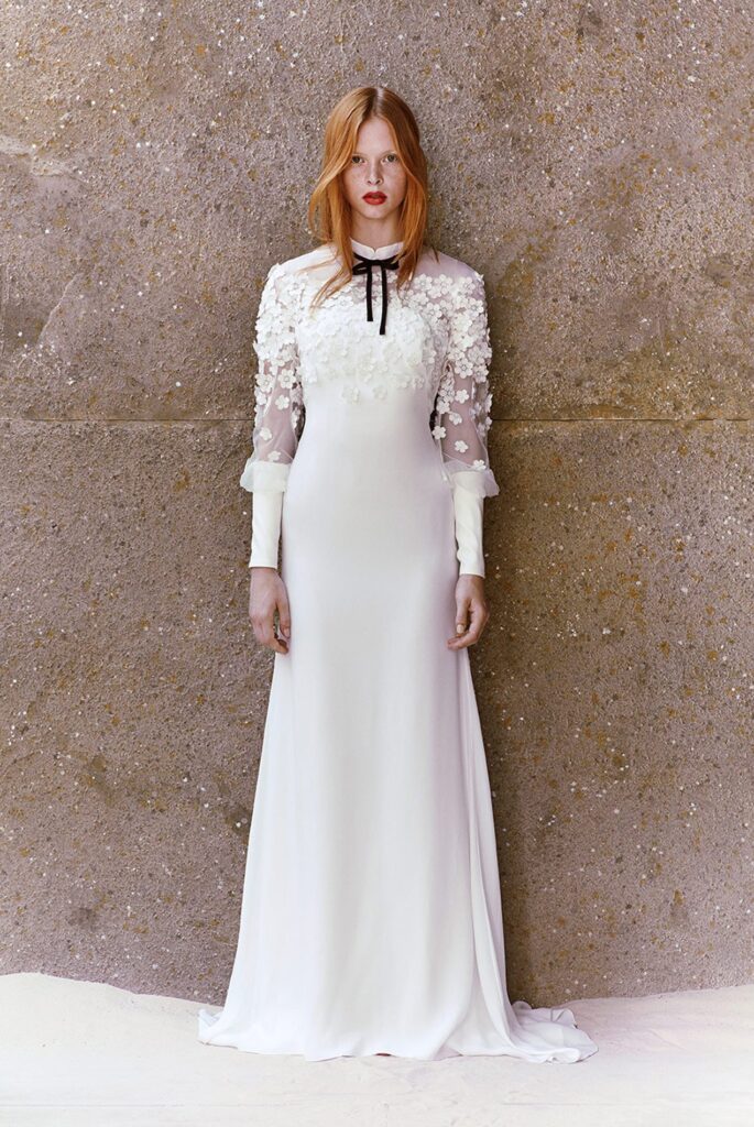 Woman in a long white dress with floral applique stands against a textured stone wall, looking at the camera.