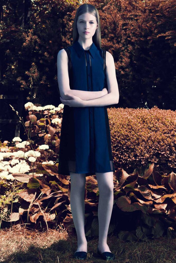 Woman in a sleeveless navy dress stands outdoors with arms crossed, surrounded by leafy plants and shrubs.