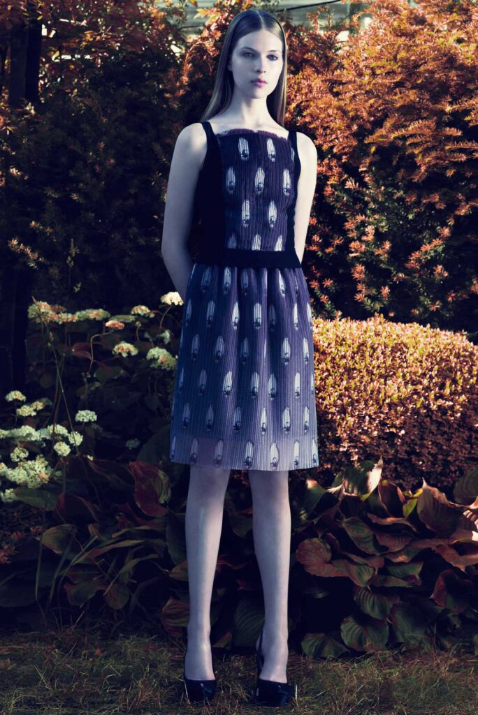 Woman in a sleeveless black and blue dress stands in a garden with lush foliage and flowers around her.