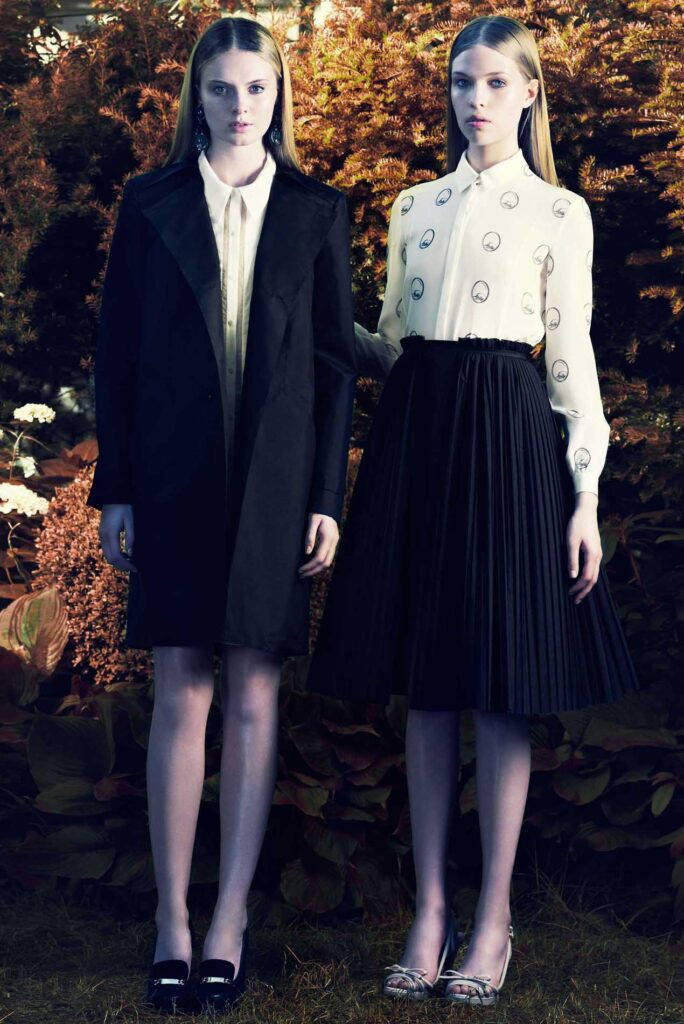 Two women stand outdoors; one wears a black coat, the other a white blouse with a black pleated skirt.