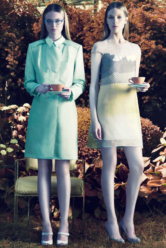 Two women in stylish dresses hold teacups while standing outdoors among bushes and flowers.
