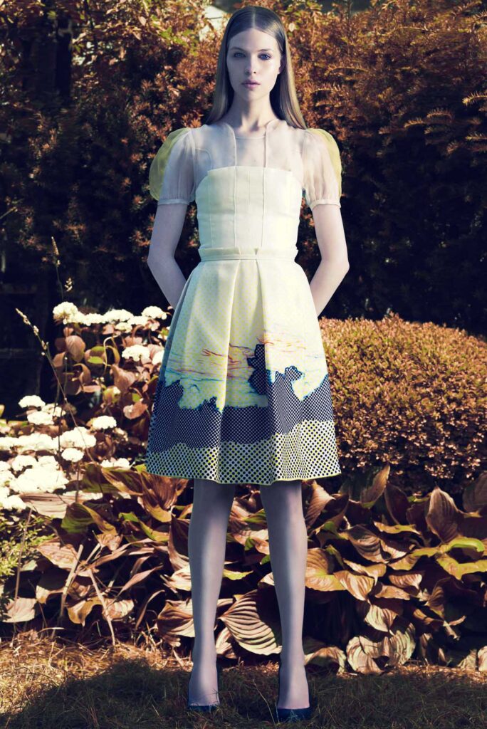 Woman in a short-sleeved white dress with artistic print stands outdoors among bushes and flowers.