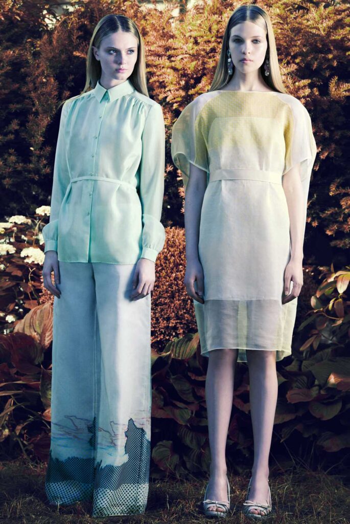 Two women stand outdoors wearing pastel sheer outfits, one in wide pants and shirt, the other in a knee-length dress.