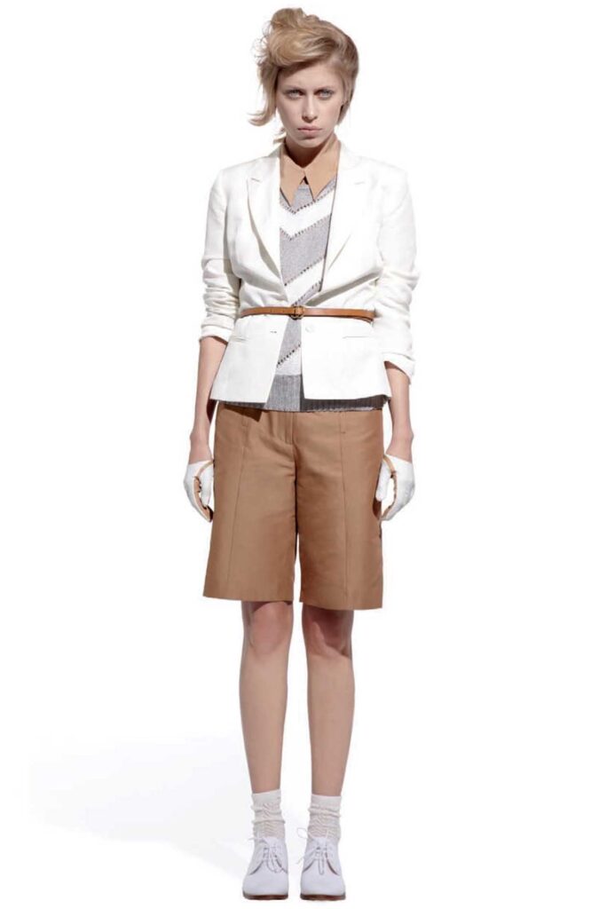 Person in a white blazer, brown shorts, striped shirt, white gloves, and brown belt, standing on white.