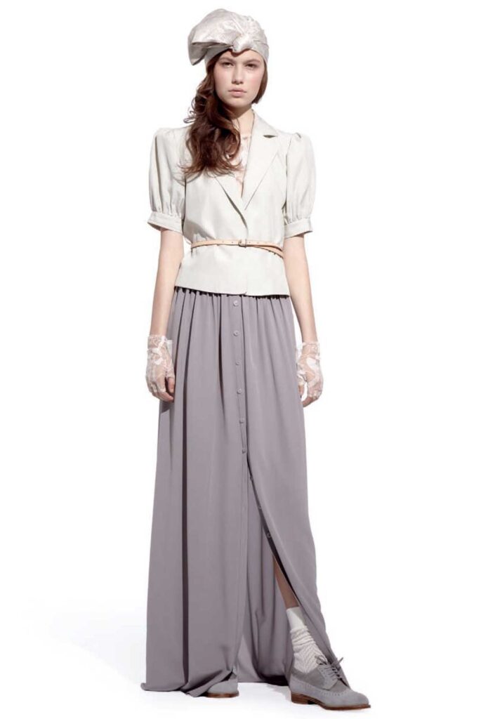 Woman in vintage-style outfit: light blouse, long gray skirt, lace gloves, headscarf, and oxford shoes.