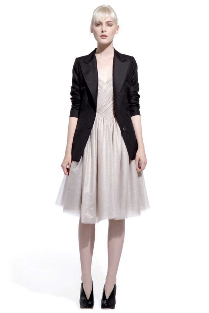 Woman in a black blazer over a light, knee-length dress, standing against a white background.