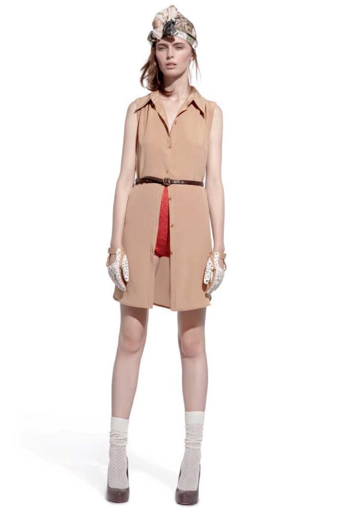Woman in a tan sleeveless dress, red shorts, gloves, headscarf, socks, and heels, standing on white background.