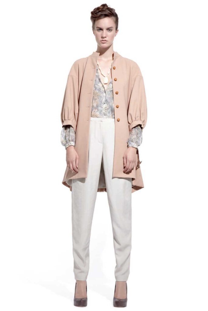 Woman in a beige coat, floral blouse, white pants, and brown heels standing against a white background.