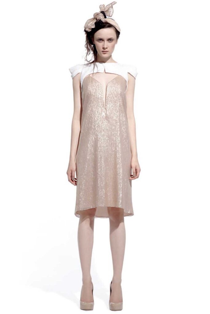 Woman in a shimmer gold dress, white cap sleeves, nude heels, and a bow headband, standing facing forward.