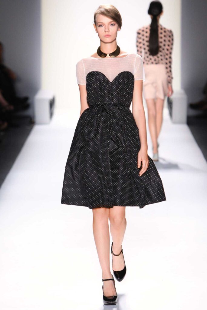 Model in a black polka dot dress and black heels walks on a white runway at a fashion show.