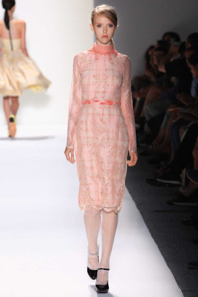 Model in a pink, long-sleeved lace dress walks down a white runway at a fashion show.