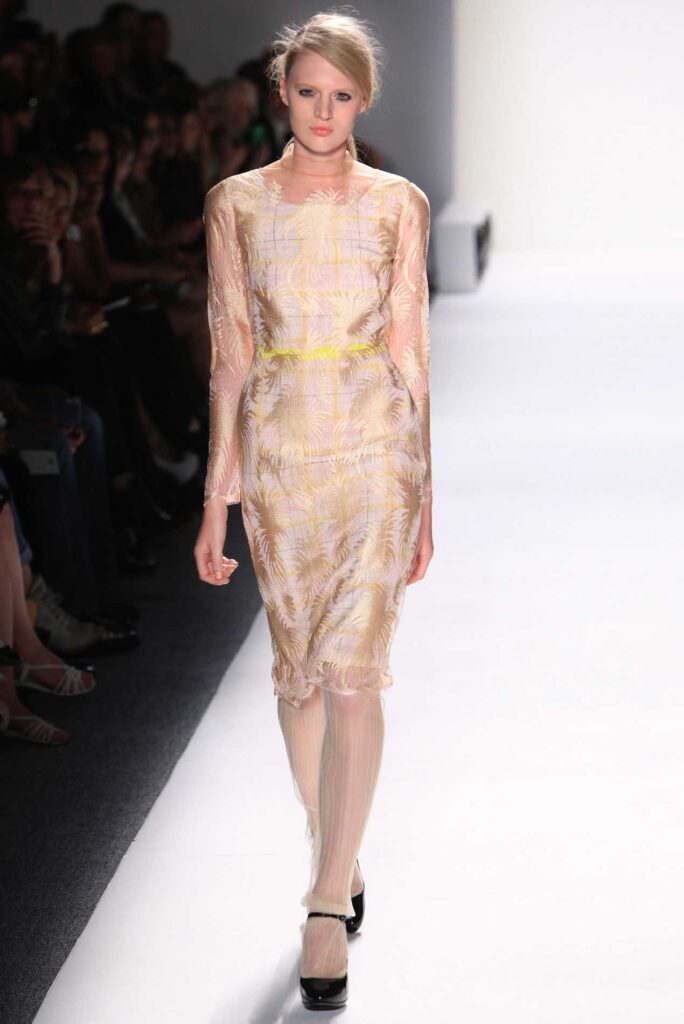 Model walks runway in sheer, long-sleeved dress with light patterns and black platform heels at a fashion show.