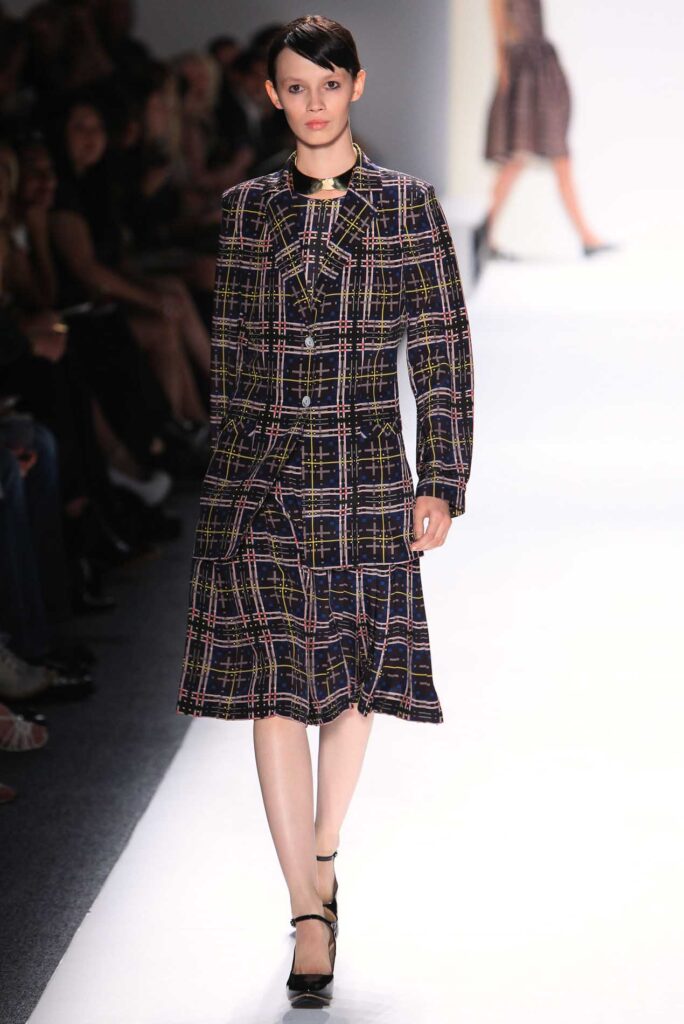 Model walks runway in a dark plaid jacket and matching skirt with ankle-strap heels at a fashion show.