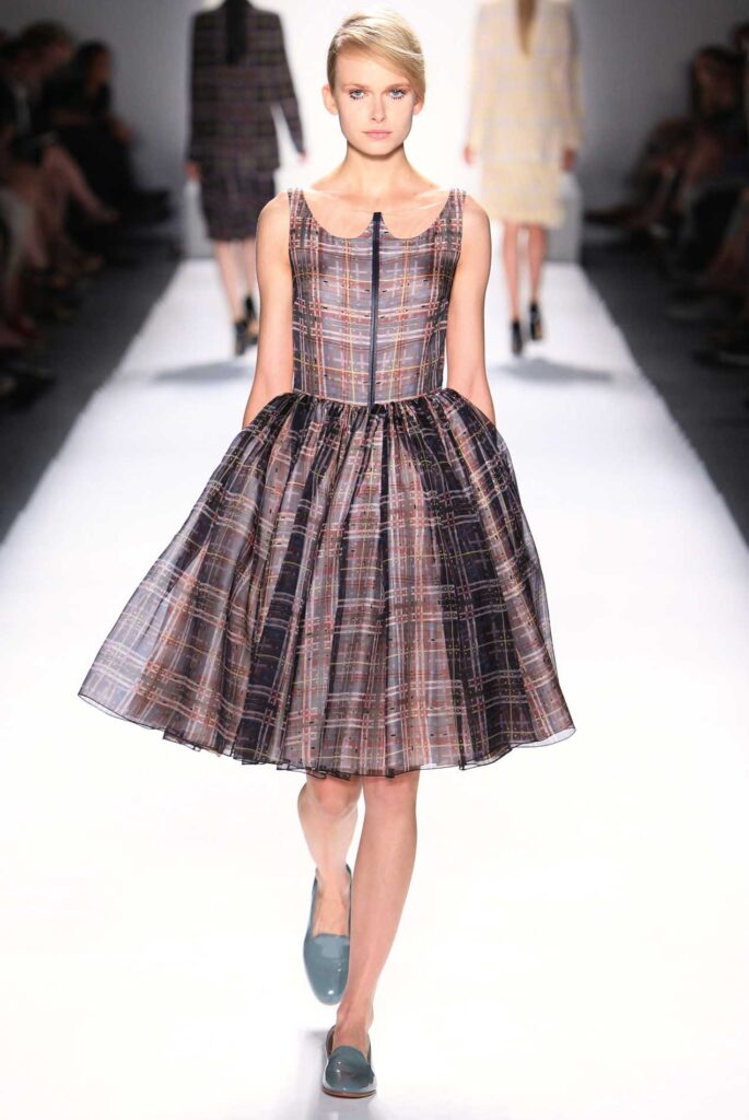 Model walks runway in sleeveless, plaid dress with full skirt and teal shoes; blurred background.