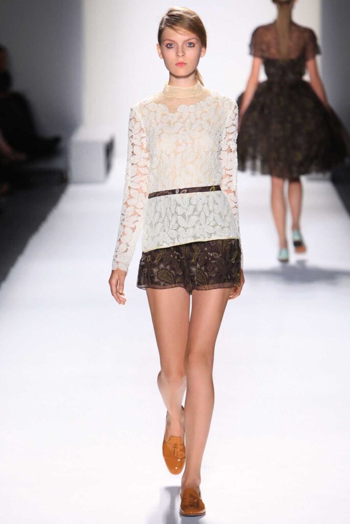 Model in a white lace top, dark floral shorts, and brown loafers walks on a fashion runway.