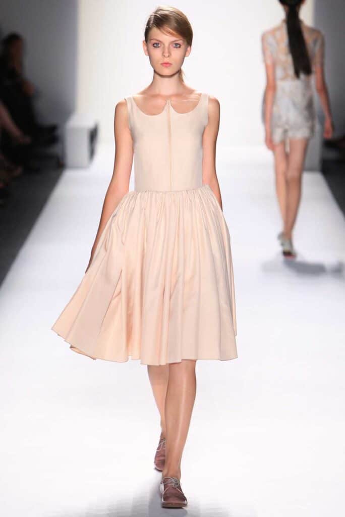 Model walks runway in a light pink sleeveless dress with a flared skirt and flat shoes.